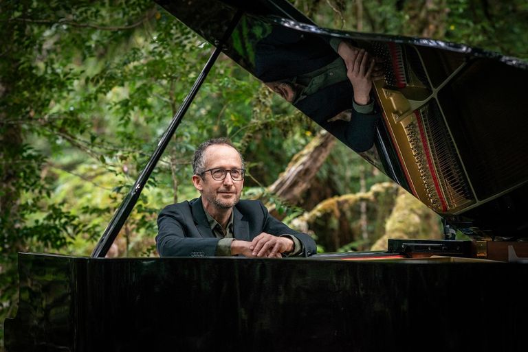 Pianist Dave McEvoy leaning on an open grand piano outdoors for Art for Takayna in a lush forest setting.